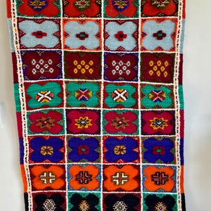 Könnte beinhalten: Ein farbenfroher, rechteckiger Wandbehang mit einem Gittermuster aus floralen Designs. Der Teppich hat eine Vielzahl von Farben, darunter Rot, Blau, Grün, Orange und Schwarz. Die Ränder sind mit weißen Fransen und einem weißen Rand versehen.