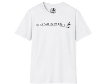 Puede incluir: Camiseta blanca con el texto negro "TO CREATE IS TO REBEL" y un dibujo de pincel. La camiseta tiene cuello redondo y mangas cortas. Un pequeño logotipo de pincel está en la parte superior de la espalda.