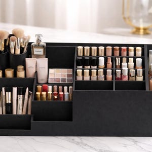 Puede incluir: Un organizador de maquillaje negro de varios niveles lleno de varios productos de belleza. Cuenta con compartimentos para brochas, pintalabios, esmaltes de uñas y productos para el cuidado de la piel. El organizador está sobre una superficie de mármol.