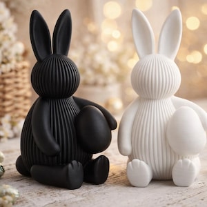 Pode incluir: Duas figuras decorativas de coelhos da Páscoa, uma preta e outra branca, cada uma segurando um ovo. Os coelhos têm detalhes verticais canelados e orelhas longas. Ovos de Páscoa e flores ao fundo.