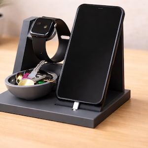 Könnte beinhalten: Dunkelgraue Ladestation mit einem schwarzen Smartphone, einer schwarzen Smartwatch und einer kleinen Schale mit Schlüsseln. Das Telefon ist angeschlossen. Die Station steht auf einer Holzoberfläche.
