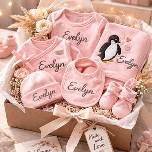 Puede incluir: Un juego de regalo rosa para bebé en una caja, que incluye un body, una camiseta cruzada, un babero, un gorro, una manta con un diseño de pingüino y botines. El nombre "Evelyn" está bordado en cada artículo. Una tarjeta dice "Welcome Baby".