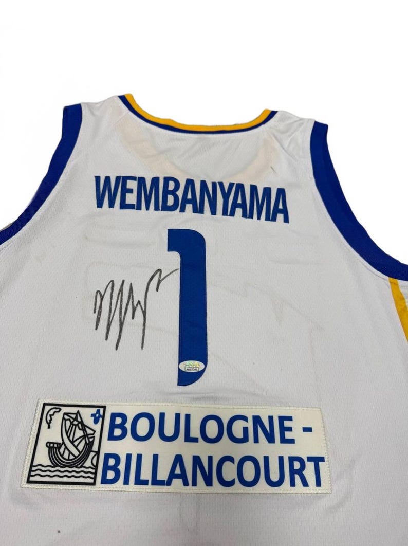 May include: White basketball jersey with blue and gold accents. The jersey features the name "WEMBANYAMA" in blue block letters, the number "1", and a signature. A patch at the bottom reads "BOULOGNE-BILLANCOURT".