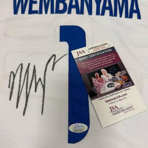 May include: White jersey with the name "WEMBANYAMA" in blue block letters. A large blue number "1" is on the front. Includes a JSA Authentication sticker and certification card. The jersey has a signature in black marker.