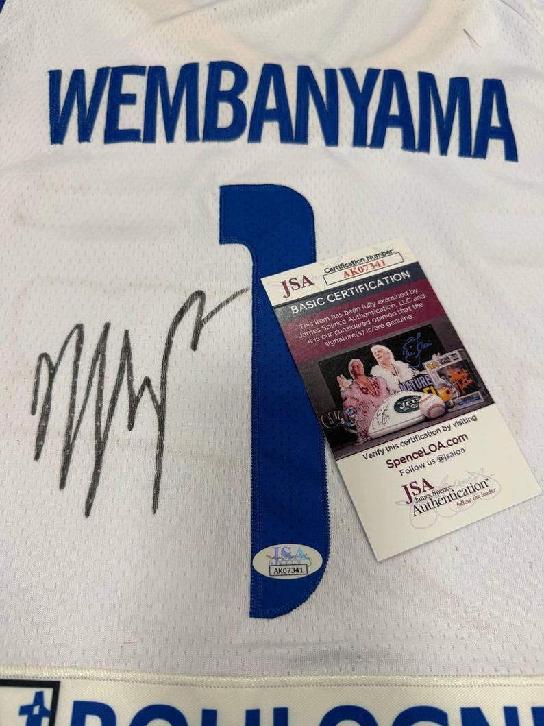 May include: White jersey with the name "WEMBANYAMA" in blue block letters. A large blue number "1" is on the front. Includes a JSA Authentication sticker and certification card. The jersey has a signature in black marker.