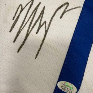 May include: White jersey with a blue stripe and a large, dark signature. A UDA authentication sticker is visible. The jersey has a textured pattern.