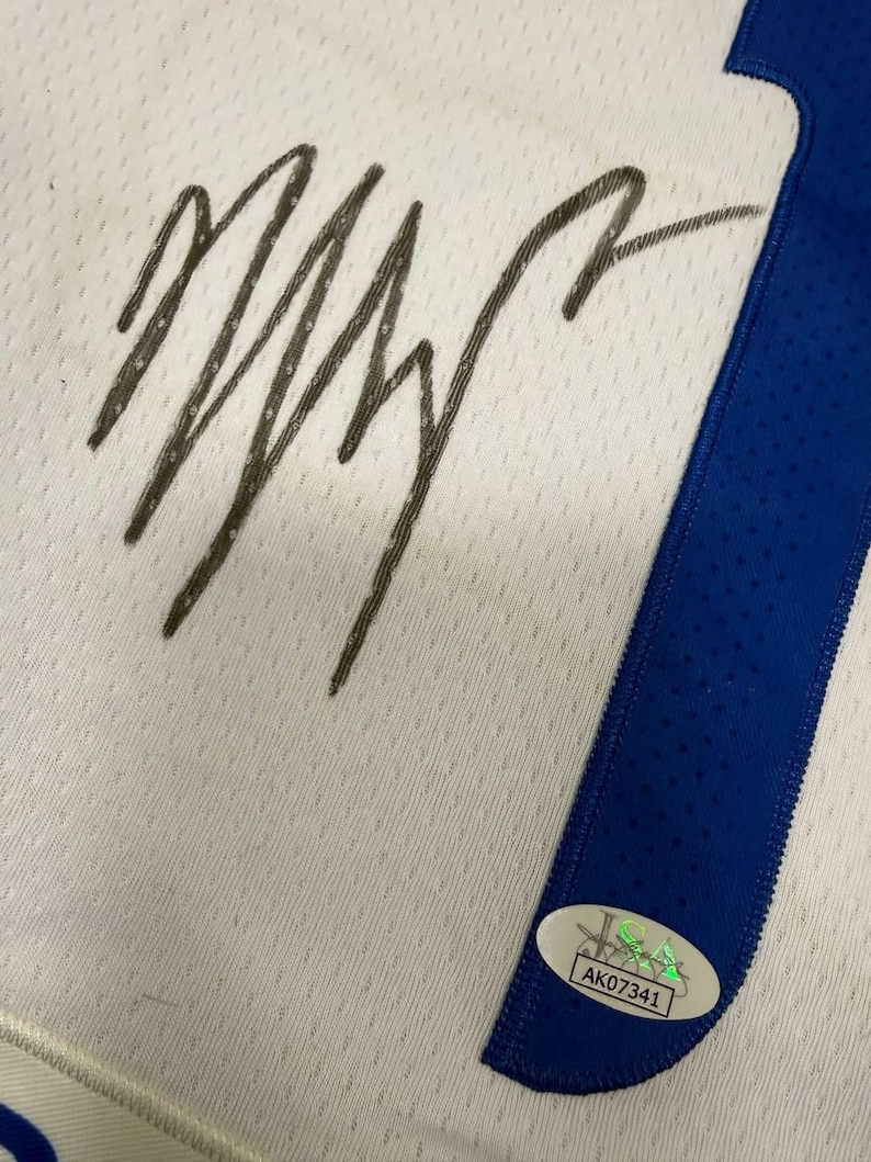 May include: White jersey with a blue stripe and a large, dark signature. A UDA authentication sticker is visible. The jersey has a textured pattern.