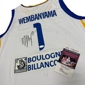 May include: White basketball jersey with blue and gold trim. The jersey features the name "WEMBANYAMA" and the number "1" in blue. Includes a "BOULOGNE BILLANCO" patch and a JSA authentication tag.
