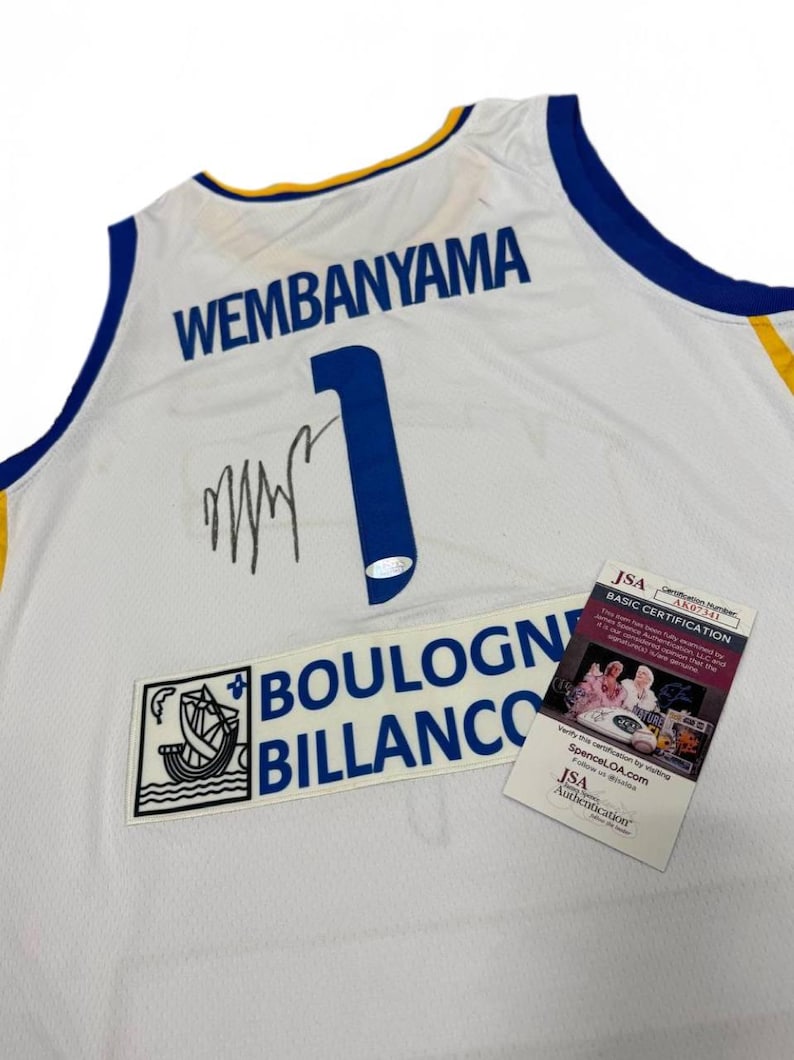 May include: White basketball jersey with blue and gold trim. The jersey features the name "WEMBANYAMA" and the number "1" in blue. Includes a "BOULOGNE BILLANCO" patch and a JSA authentication tag.