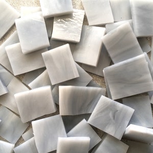 May include: A pile of small, square, gray glass tiles with a subtle marbled pattern. The tiles are perfect for crafting and mosaic projects.