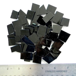 May include: A pile of black square tiles, measuring approximately 1 inch by 1 inch, are scattered on a white surface. A ruler is visible in the bottom of the image, showing the size of the tiles.