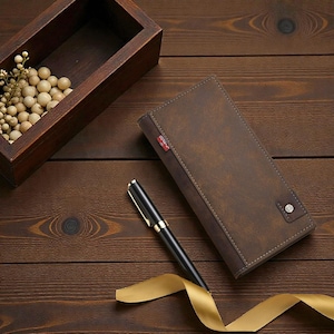 May include: A brown leather wallet with a button closure and a small red tag. A black pen with gold accents and a gold ribbon are next to the wallet. A wooden box with decorative spheres is in the background.