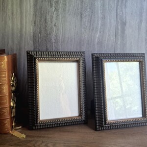 May include: Two rectangular picture frames with dark, textured, ridged borders and inner gold-toned trim. The frames are empty, featuring clear glass fronts, and are positioned upright next to a stack of three leather-bound books with gold lettering.