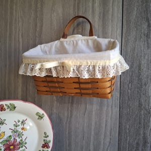May include: A woven brown basket with a white fabric lining and a brown leather handle. The basket has a cream-colored lace trim and is hanging on a gray wall. A decorative plate with floral and bird designs is in the foreground.
