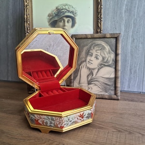 May include: An octagonal jewelry box with a gold-toned frame and a hinged lid featuring an interior mirror. The exterior base is decorated with a floral pattern in shades of red, orange, and grey. The interior is lined with vibrant red velvet.