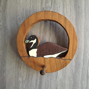 May include: Wooden wall hanging featuring a carved duck within a circular frame. The duck has a black head, white cheek markings, and brown body. The frame is made of light brown wood and includes a small wooden knob at the bottom.