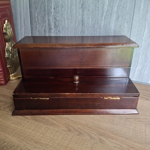 May include: A dark brown wooden desk organizer with a tiered design. The unit features a raised back section and a lower front compartment with a small round knob and two visible gold-toned metal hinges on the lid, showcasing a smooth finish.