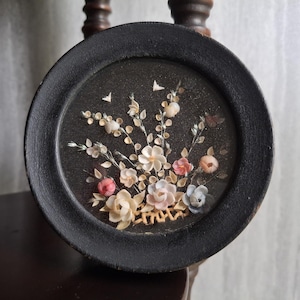 May include: A round, black framed decorative piece featuring a floral arrangement made of seashells. The arrangement includes white, pink, and blue flowers, with small shell butterflies. The background is dark.