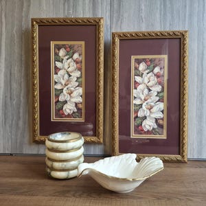 May include: Two framed floral prints with gold frames and a white flower arrangement. A stack of marble coasters with gold trim and a white, shell-shaped dish with gold trim are in front of the prints.