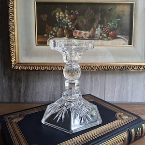 May include: A clear, cut-glass candlestick holder with a square base and a round, beaded center. The candlestick is displayed on a dark blue book with gold lettering, in front of a framed painting.