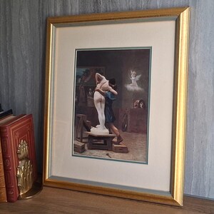May include: A framed print with a gold-colored frame. The artwork depicts a sculpture of a nude figure being embraced by a man. The print is displayed on a wooden surface next to a stack of books and a decorative bookend.