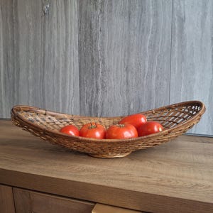 May include: A woven wicker basket in a boat shape, filled with ripe red tomatoes. The basket sits on a wooden surface against a gray wall. The tomatoes are a vibrant red color.