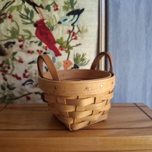 May include: A small, round, woven wooden basket featuring a light tan finish, two sturdy leather handles attached to the sides, and a reinforced upper rim secured with small metal rivets, showcasing a classic splint-style construction.