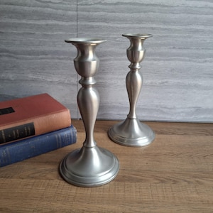 May include: Two silver-colored candle holders with a classic design. The candle holders are placed on a wooden surface, with two antique books in the background. The candle holders have a wide base and a flared top.