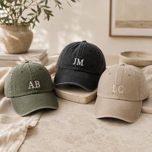 Può includere: Tre cappellini da baseball in verde oliva, nero antracite e beige. Ogni cappellino presenta una struttura a sei pannelli con visiera curva e iniziali bianche ricamate sul davanti: AB sul verde, JM sul nero e LC sul cappellino color beige.