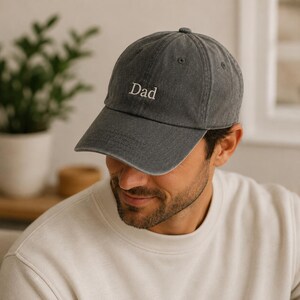 Puede incluir: Una gorra de béisbol gris carbón con visera curva y el texto Dad bordado en blanco centrado en el panel frontal. La gorra presenta una construcción clásica de seis paneles con ojales cosidos y un diseño de corona estructurada.
