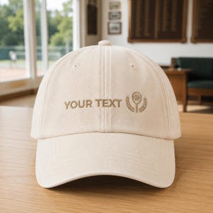 Può includere: Un cappellino da baseball beige con il testo ricamato "YOUR TEXT" in marrone, accompagnato da una piccola icona marrone di una racchetta da pickleball circondata da un disegno a corona d'alloro, centrato sul pannello frontale del cappellino.