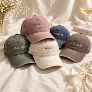 May include: A collection of five baseball caps in muted colours including dusty rose, olive green, cream, navy blue, and brown. Each cap features embroidered white text reading Bride, Maid of Honor, Wifey, Team Bride, and Bridesmaid respectively.