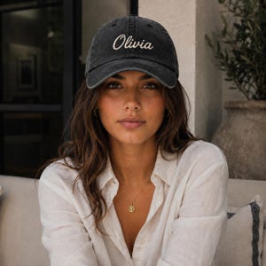Op de afbeelding: Een antracietgrijze honkbalpet met een verwassen afwerking. Op de voorkant staat de naam Olivia geborduurd in een wit, cursief lettertype. De pet heeft een gestructureerde kroon, een gebogen klep en een knoopje aan de bovenkant.