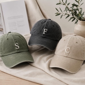 Op de afbeelding: Drie baseballpetten in olijfgroen, antracietgrijs en beige. Elke pet heeft een enkele geborduurde witte letter op het voorpaneel: een S op de olijfgroene pet, een F op de antracietgrijze pet en een C op de beige pet. De petten hebben een gebogen klep.