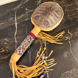 May include: A handcrafted Native American rattle with a round, patterned head and a beaded handle. The handle is wrapped with colourful beads and leather fringe. The rattle features small metal tubes and colourful beads.