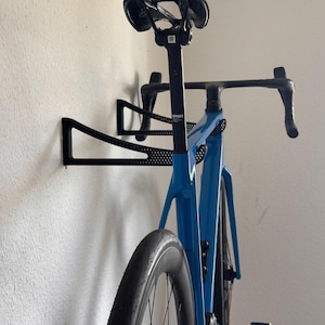 Könnte beinhalten: Ein blaues Rennrad, das an einem schwarzen Wandfahrradträger befestigt ist. Das Fahrrad hat einen schwarzen Sattel, Lenker und Räder. Der Träger besteht aus schwarzem Metall und ist an einer weißen Wand befestigt.