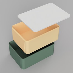 Könnte beinhalten: Dreistufige, minimalistische Aufbewahrungsbehälter in einem Stapel. Der untere Behälter ist ein gedämpftes Grün, der mittlere ist ein helles Beige und die Oberseite ist ein weißer Deckel. Die Behälter sind rechteckig und scheinen aus einem festen Material zu bestehen.