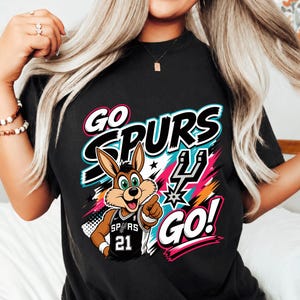 May include: Black t-shirt with a cartoon jackrabbit wearing a Spurs jersey, the text "GO SPURS GO!" in white, teal, and pink. The jackrabbit is pointing with its right hand.