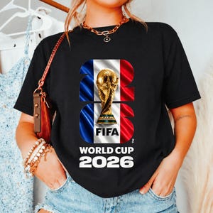 May include: A black short-sleeve t-shirt featuring a graphic of the FIFA World Cup 2026 trophy centered over a blue, white, and red vertical striped background. The text 2026 is integrated into the design, with World Cup 2026 printed below.