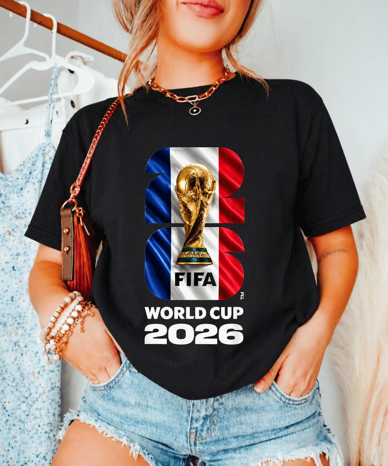 May include: A black short-sleeve t-shirt featuring a graphic of the FIFA World Cup 2026 trophy centered over a blue, white, and red vertical striped background. The text 2026 is integrated into the design, with World Cup 2026 printed below.