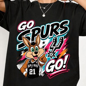 May include: Black t-shirt featuring a cartoon jackrabbit wearing a Spurs jersey with the number 21. The shirt has the words "GO SPURS GO!" in white, black, pink, and teal. A colorful graphic design is behind the jackrabbit.