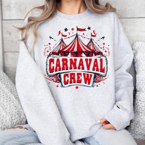 Puede incluir: Sudadera gris claro con un diseño de temática de carnaval en rojo, blanco y azul. El diseño presenta carpas de circo, banderas, estrellas y las palabras "CARNAVAL CREW". Es una prenda informal de manga larga.