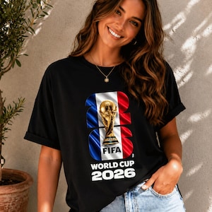 May include: A black short-sleeved t-shirt featuring a graphic of the FIFA World Cup 2026 trophy centered over a vertical blue, white, and red flag design. The text FIFA WORLD CUP 2026 is printed in white below the trophy and flag graphic.