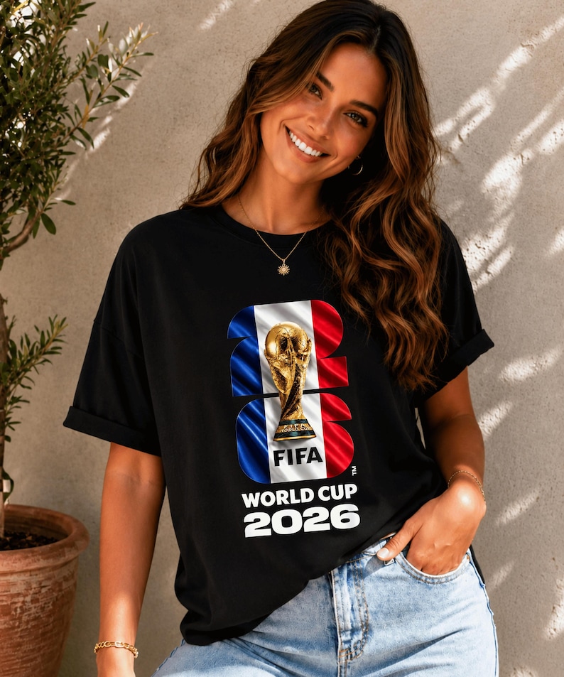May include: A black short-sleeved t-shirt featuring a graphic of the FIFA World Cup 2026 trophy centered over a vertical blue, white, and red flag design. The text FIFA WORLD CUP 2026 is printed in white below the trophy and flag graphic.
