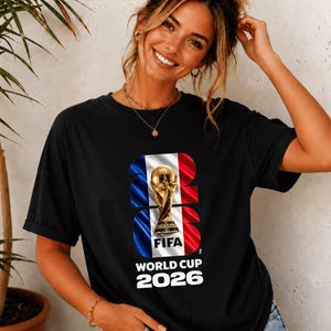 May include: A black short-sleeved t-shirt featuring a graphic print of the FIFA World Cup 2026 trophy centered over a vertical blue, white, and red flag design. The text 2026 is integrated into the graphic, with World Cup 2026 printed below.