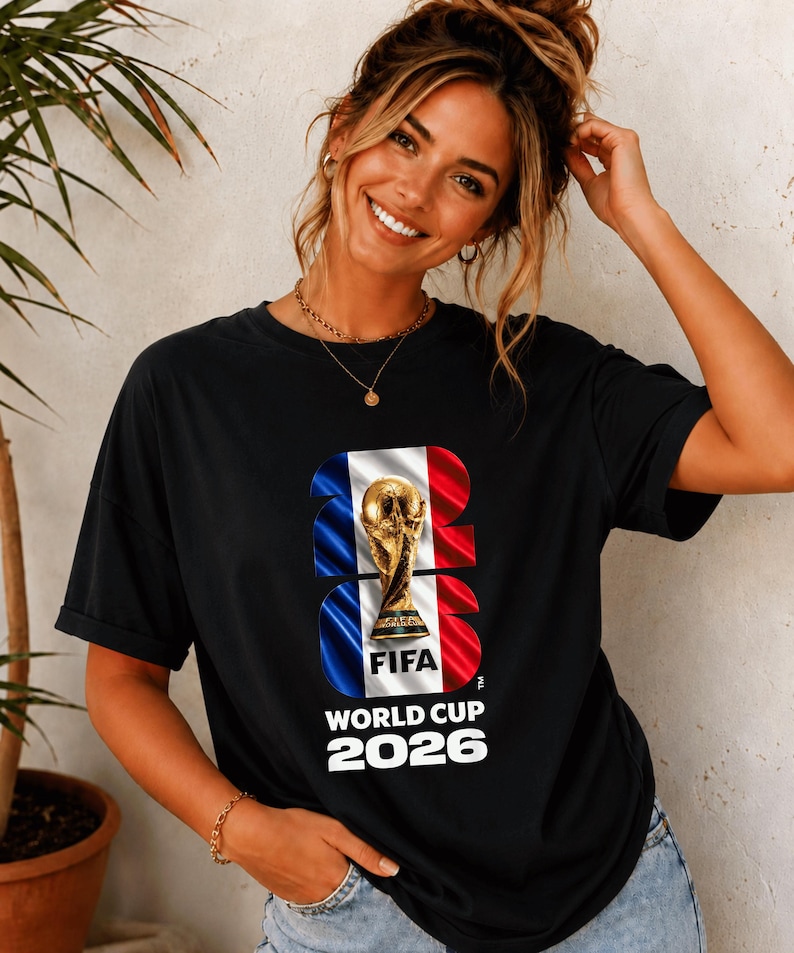 May include: A black short-sleeved t-shirt featuring a graphic print of the FIFA World Cup 2026 trophy centered over a vertical blue, white, and red flag design. The text 2026 is integrated into the graphic, with World Cup 2026 printed below.