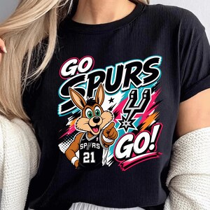 May include: Black t-shirt featuring a cartoon jackrabbit wearing a Spurs jersey with the number 21. The shirt has the words "GO SPURS GO!" in a colorful design with teal, pink, and orange accents. The jackrabbit is pointing.