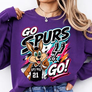 May include: A purple sweatshirt with a graphic featuring the words "GO SPURS GO!" and a cartoon jackrabbit wearing a basketball jersey with the number 21. The design includes black, white, teal, pink, and orange accents.
