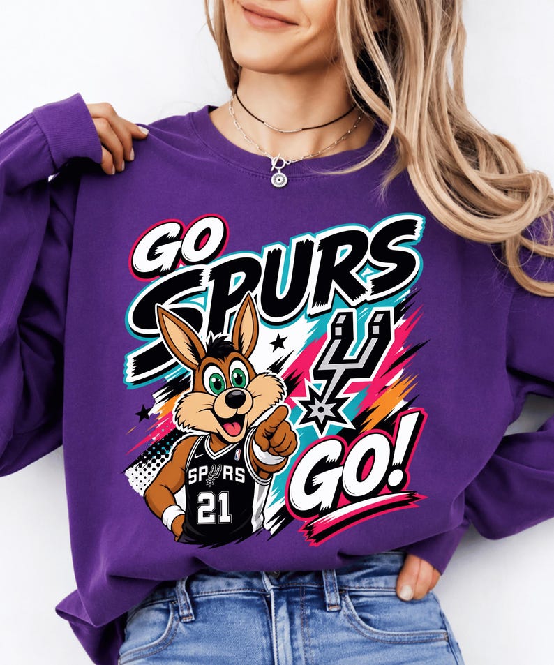 May include: A purple sweatshirt with a graphic featuring the words "GO SPURS GO!" and a cartoon jackrabbit wearing a basketball jersey with the number 21. The design includes black, white, teal, pink, and orange accents.