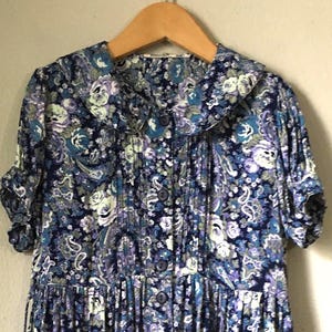 May include: A vintage floral print dress with a Peter Pan collar. The dress features a navy blue background with a paisley pattern in shades of white, light blue, and purple. It has short sleeves and is hanging on a wooden hanger.
